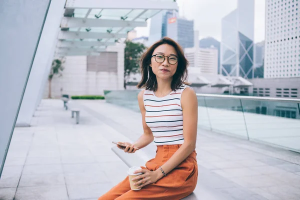 香港のカメラを見ながら ガラスの中で笑顔とプラスチック製のカップとスマートフォンでベンチに座って美しい若いアジアの女性 — ストック写真