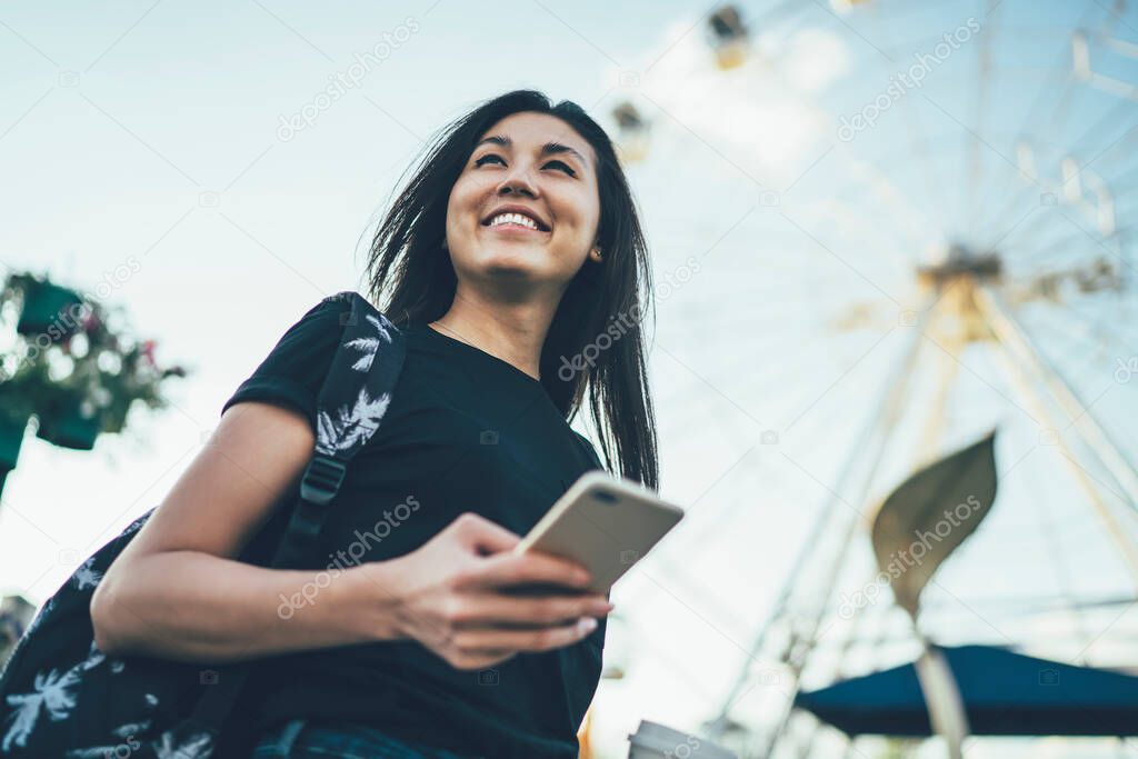 Below view of attractive smiling woman with cellular gadget in hands standing outdoors and laughing feeling good mood from spending day in Amusement Park, happy hipster girl with smartphone