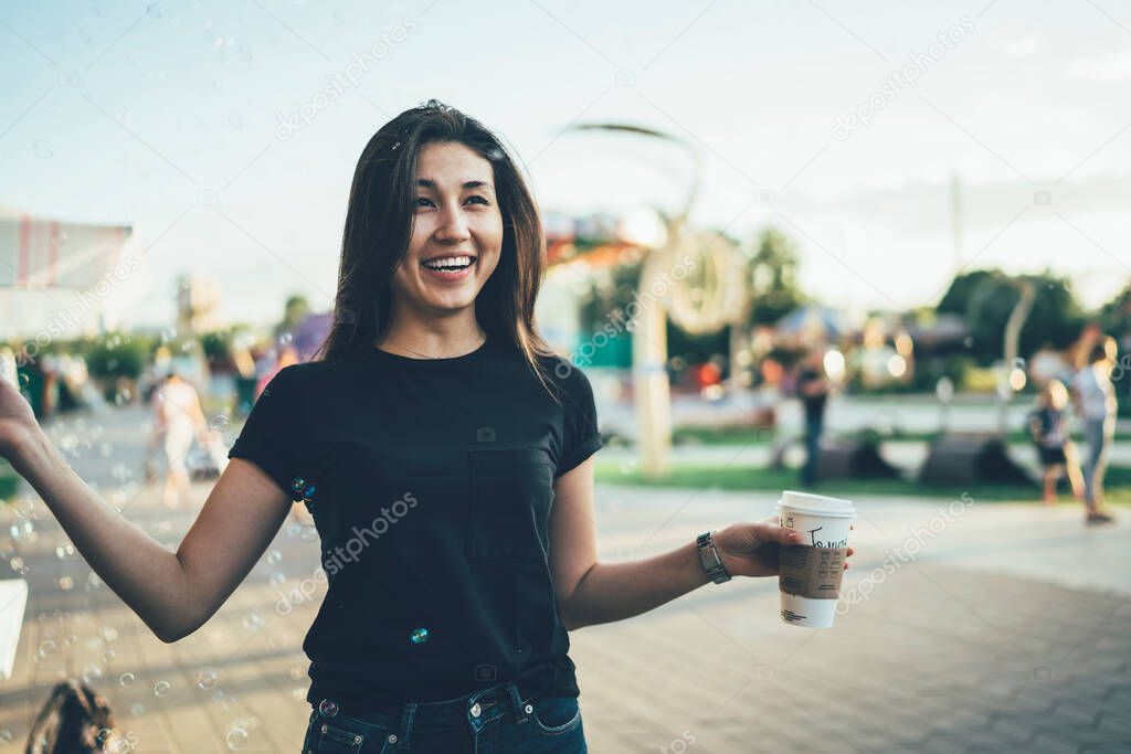 Happy woman in blank t-shirt holding takeaway cup in hand and rejoicing during free time in Amusement Park, cheerful Asian hipster girl feeling playful mood during recreation day in public place