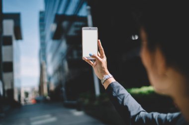 Siyah ceketli genç bir kadın güneşli bir günde şehir merkezinde cep telefonuyla fotoğraf çekiyor.