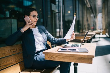 Kafe masasında uzaktan kumandalı cihazlarla çalışırken iş kâğıtlarına dikkatle bakan dalmış sakin bir kadın.