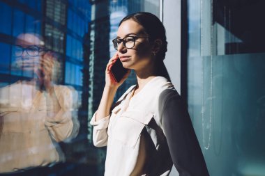 Beyaz bluzlu hırslı genç bir girişimci iş yerindeki pencereden dışarı bakarken cep telefonuyla konuşuyor. 