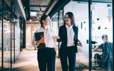 Resmi tarzda giyinen içerik siyah saçlı bayan meslektaşlar modern ofis binasında konuşurken ve kafeye gagalarıyla giderken gülümserler.