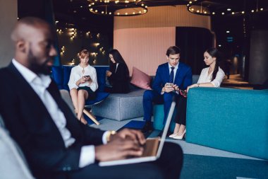 Çalışan genç dalgın iş ekibi, iş sonrası konferans sırasında lobideki kanepe ve koltuklarda ayrı ayrı dinlenirken cihazlara göz atıyorlar.