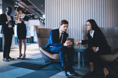 Rahat koltuklarda oturan ve dijital tabletle etkileşime geçen düşünceli, kendini işine adamış iş adamları modern açık uzay ofisinde ayakta durup akıllı telefon kullanarak eğlenceli sohbetler yapıyorlar.