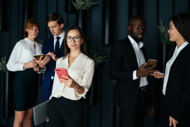 Düşük açılı, kendine güvenen sakin iş kadını ve dizüstü bilgisayarlı resmi kıyafetler kameraya bakarken akıllı telefon kullanarak düşünceli yetişkin çok ırklı iş arkadaşlarının konferans sonrası ofis koridorlarında dijital cihazlarla etkileşim kurması.