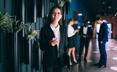 Resmi takım elbiseli mutlu genç bir kadın elinde evrak işleriyle arkadaşlarıyla mesajlaşarak modern ofis alanında kameralara bakarak vakit geçiriyor.