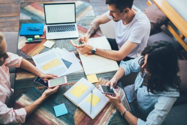 Yüksek açılı genç iş arkadaşları açık not defterleri ve boş ekranlı dizüstü bilgisayarın yanında renkli kağıtlarla masada otururken akıllı telefonlarda mesajlaşıyorlar.