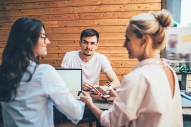 Akıllı telefonlu genç, gülümseyen sarışın kadın dizüstü bilgisayarlı kadın esmer meslektaşına bakarken, erkek partner kameraya ilgiyle bakıyor.