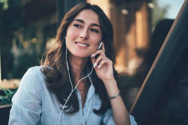 Uzun saçlı, siyah saçlı, mavi tişörtlü ve kafede şarkı dinlemek için beyaz kulaklıklı genç bir kadın. 