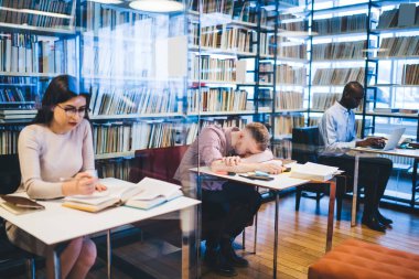 Çok ırklı girişimciler resmi kıyafetler giyip kitap okuyor ve kütüphanede yüzüstü yatan yorgun iş arkadaşının yanında aletler kullanıyor.
