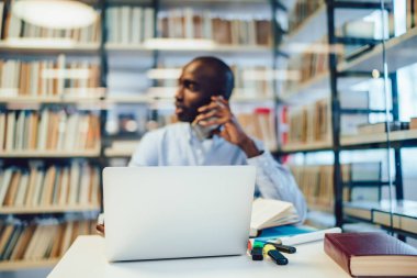 Çeşitli kırtasiye ve modern dizüstü bilgisayar kütüphane masasının yanına yerleştirilmiş. Bulanık Afrikalı Amerikalı bir adam akıllı telefondan kitaplığın yanında konuşuyor.