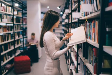 Kitap rafının önünde duran ve elinde kitap tutan güzel, ince bir kadının yan görüntüsü. 