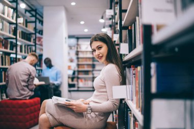 Çekici ve neşeli bir kadının kütüphanede bacak bacak bacak üstüne atmış kitap rafına yaslanmış, elinde kitap tutarken kameraya bakarken görüntüsü.