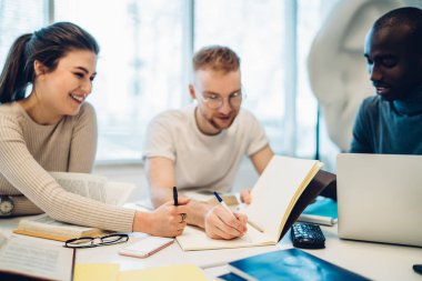 Modern kütüphanede kitaplarla ve dizüstü bilgisayarlarla masada otururken gündelik giysiler içinde özenli bir şekilde gülümseyen çok ırklı gençler deftere yazıyorlar.