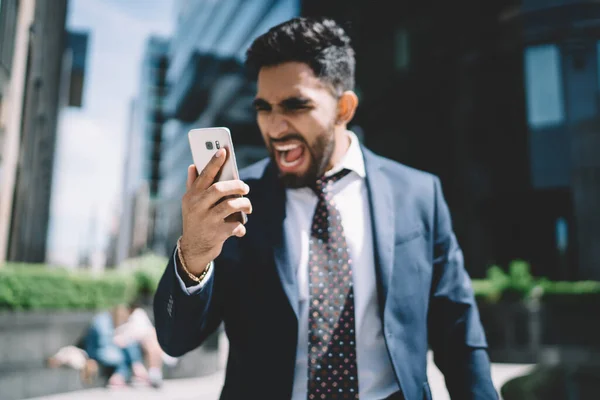 Adult mad bearded businessman wearing formal clothing looking at screen and reading message on cellphone while screaming loud with mouth open. Stressed furious executive shouting during call on phone