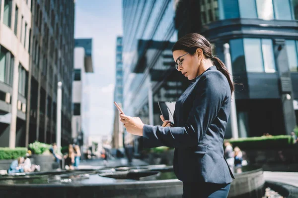 現代都市の路上で同僚と電話通信を使用して手に文書を持つエレガントなスーツと眼鏡の普及した深刻な女性の側面図 — ストック写真
