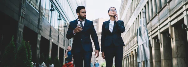 Smart serious business man and woman in classical elegant suit walking on street and speaking on mobile phone on way in business center of downtown
