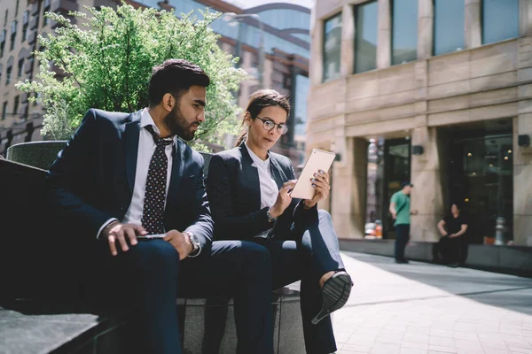 Focused woman in formal wear and glasses with crossed legs using tablet relaxing from work with partner sitting on fence in street of modern city