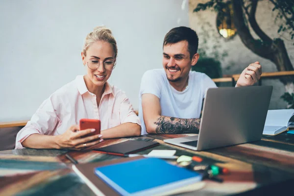 Tattooed and bearded brunet guy and blond young woman sitting at table with laptop and notepads sharing smartphone and having fun