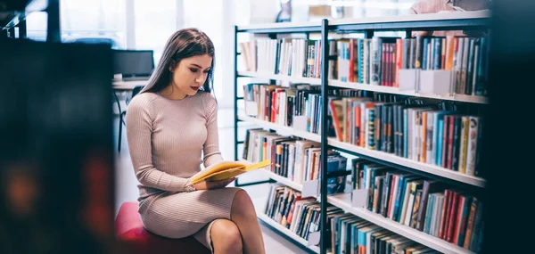穿着时髦衣服的年轻貌美的女学生坐在书架旁 在现代大学图书馆读书 同时阅读有趣的书籍 — 图库照片