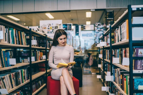 Beautiful young woman in beige dress sitting on pouf and reading book with yellow cover while sitting near bookshelves in university library