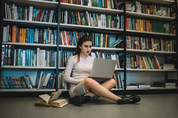 Concentrated confident student woman studying with laptop and open book sitting with crossed legs on floor leaning on bookcase in light library