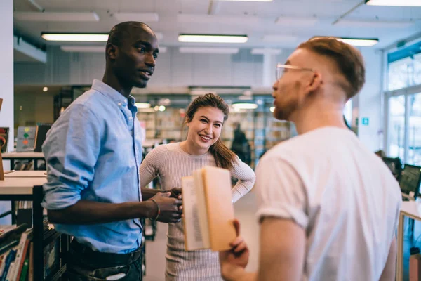 Optimistic and carefree female student standing in library and smiling while other members of multiethnic study group arguing to each other