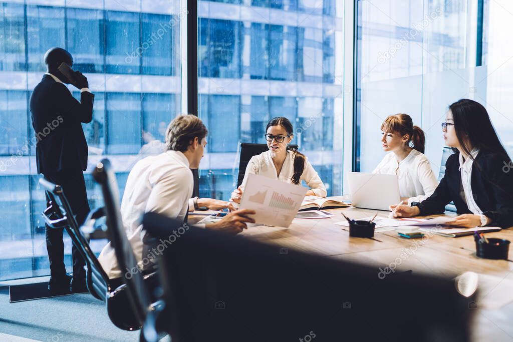 Group of young multiracial coworkers having meeting at conference room and sharing opinions about financial results while male colleague standing at window and talking on phone