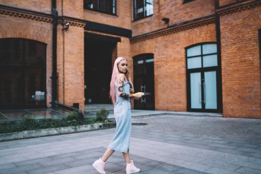 Dövmeli, uzun pembe saçlı, kot elbiseli bir kadın. Cep telefonuyla konuşuyor. Tuğla binayla sokakta yürüyor.