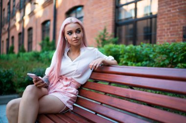 Beyaz tişörtlü, pembe şortlu, uzun pembe saçlı, cep telefonuyla müzik dinleyen caddedeki ahşap bankta oturan sıska kadın.