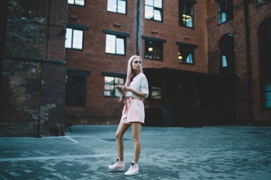 Beyaz tişörtlü, pembe şortlu, uzun pembe saçlı, cep telefonuyla mesajlaşan, caddedeki tuğla binanın karşısında duran zayıf bir kadın.