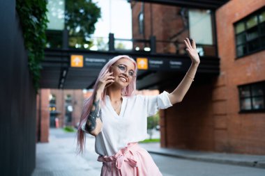 Moda gözlüklü, şık, parlak elbiseli genç, mutlu bir bayan. Şehirde telefonla konuşurken artistik bir şekilde el kaldırıyor ve başka tarafa bakıyor.