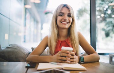 Mutlu sarışın kadın, bir bardak içkiyle masaya oturup dinlenirken gülümseyecek ve kameraya bakacak.
