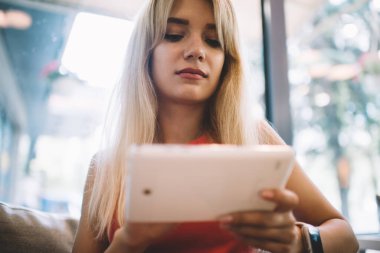 Düşük açılı, sarı saçlı, tablet kullanan, pencereye yaslanmış, rahat, modern bir kafede dinlenen bir kadın.