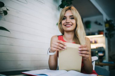 Aşağıdan mutlu, genç, çekici sarışın kadın çalışan kameraya bakıyor ve modern ofiste çalışırken ve otururken zarfı tutuyor.