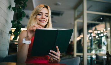 Siyah kitap okuyan ve kafede otururken ilginç komik hikayelerin tadını çıkaran neşeli, modern sarışın bir kadın.