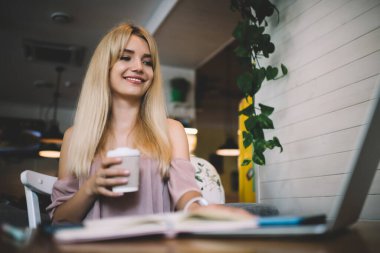 Alçak açılı, gülümseyen, açık pembe elbiseli, dizüstü bilgisayarda çalışan ve kahve dükkanının masasında bir fincan kahve tutan sarışın.