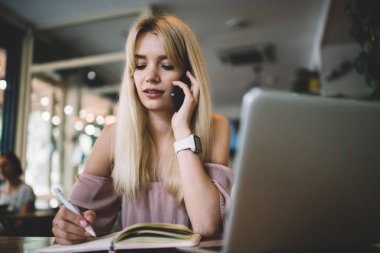 Modern saat ve pembe elbise giyen genç bir bayan kahve dükkanında telefonla konuşurken günlük planlamacıyla yazıyor. 