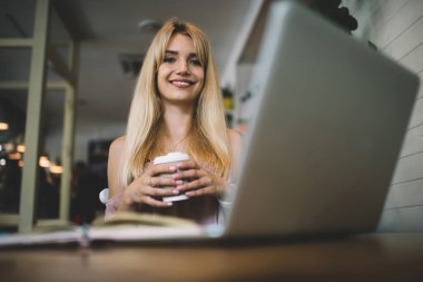 Uzun sarı saçlı, kolsuz elbiseli, kahve dükkanında oturan, elinde kahve fincanı olan ve kameraya gülümseyen bir kadın.