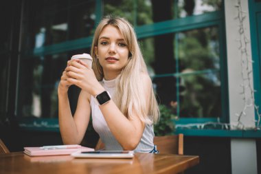 Beyaz kolsuz balıkçı yaka giyen, modern kol saati takan, kahve dükkanında elinde kahve fincanıyla oturan ve kameraya bakan sarışın genç bir kadın.