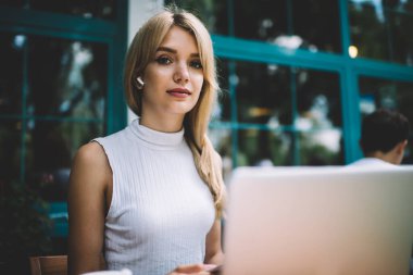 Kolsuz tişörtlü zeki sarışın kadın şehirde bir kafede dizüstü bilgisayarla oturuyor.