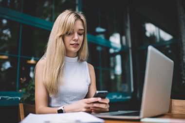 Kolsuz balıkçı yaka kazak giymiş, elinde akıllı bir telefon tutan ve gündüz kahve dükkanının önünde dizüstü bilgisayarla oturan genç sarışın bir kadın.