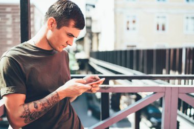 Dövmeli, gündelik kıyafetli genç bir adam cep telefonu kullanıyor. Arkadaşlarıyla mesajlaşıp sohbet ediyor. Kafe 'nin dışında, parlak güneşin altında dinleniyor.