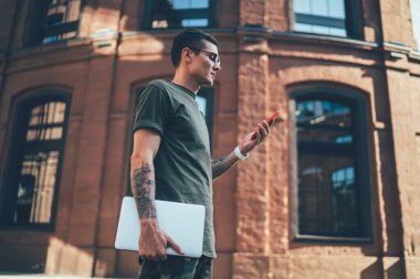 Modern netbook 'ta çalışan yetenekli bir serbest çalışanın iş birliği yapması ve toplantının zamanını kontrol etmesi için cep telefonundan gönderilen mesajlar ve internet bağlantısı olan yakışıklı bir adamın yan görüntüsü.