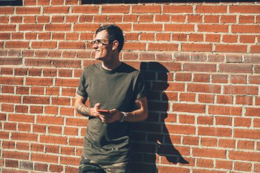 Gözlüklü ve haki renk elbiseli neşeli genç adam güneş ışığında kırmızı tuğla duvarın yanında dikilirken gözlerini kaçırıp cep telefonuyla sörf yapıyor.