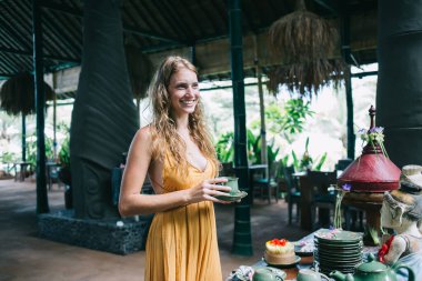 Oldukça neşeli, sarışın, turuncu elbiseli, uzun boyunlu, çay fincanı ve çay tabağı tutan genç bir kadın Bali restoranında zümrüt çay takımıyla masanın önünde dikilirken.