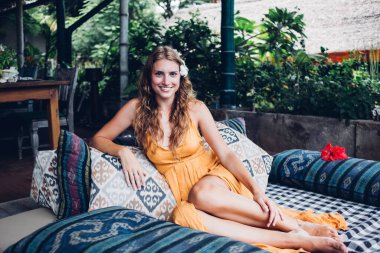 Mutlu, çıplak ayaklı, sarışın, saçları çiçekli, turuncu desenli elbisesi olan, koltukta oturan, yerel bir restoranın terasında dinlenirken dirilmiş, dirseği süs yastığına takılmış, güzel bir kadın.