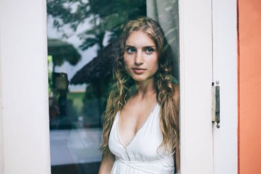 Yalnız, melankolik, beyaz elbiseli sarışın bir kadın, güneşli bir günde Bali tatil köyündeki tropik bahçeye karşı pencerenin yanında dikilirken dışarı bakıyor ve düşünüyor.