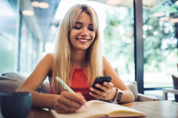 Szczęśliwa Młoda Kobieta Blond Włosy Przeglądanie Smartfona Pisanie Notebooku Podczas — Zdjęcie stockowe